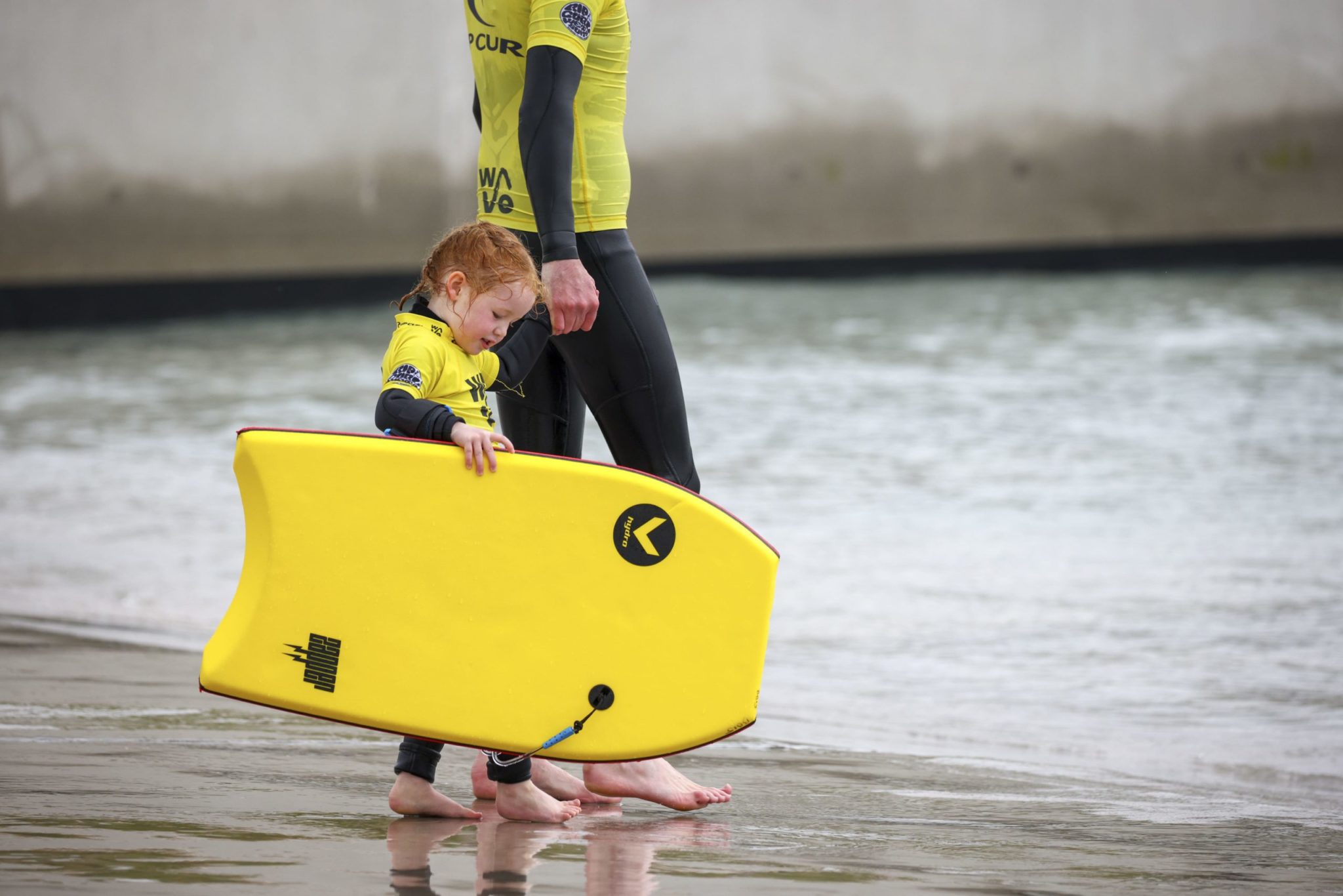 A child holding her fathers hand walking into the beginner bay with a bodyboard at The Wave bristol