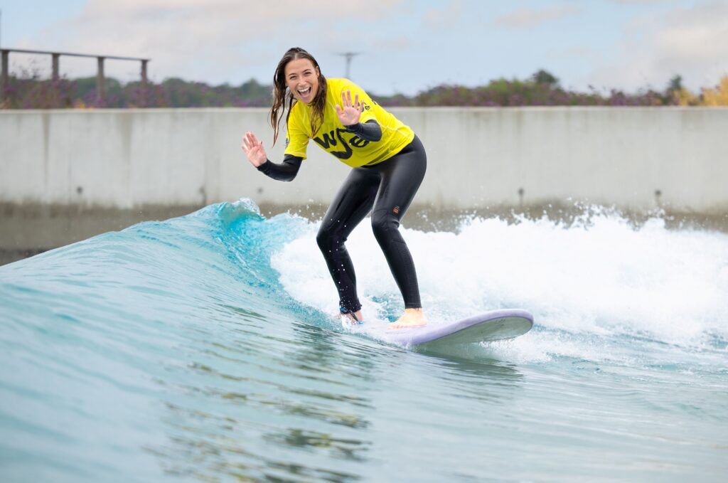 A woman smiles and waves while surfing in an Improver session at The Wave Bristol.