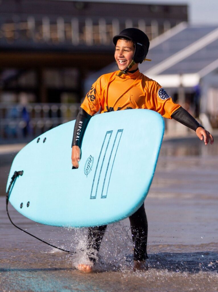 A child holds a softboard while standing on the shore at The Wave Bristol.